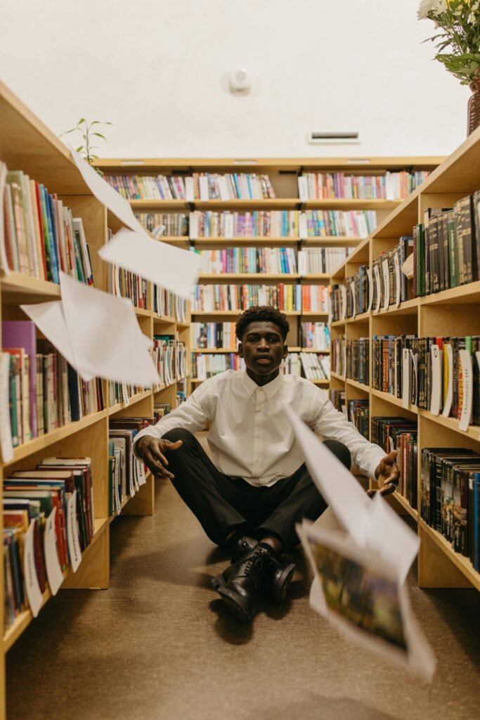 A young man sits on the library floor with papers flying around him between wooden book shelves.