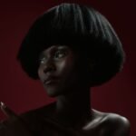 A striking studio portrait of a woman with a bold hairstyle on a dark red background.