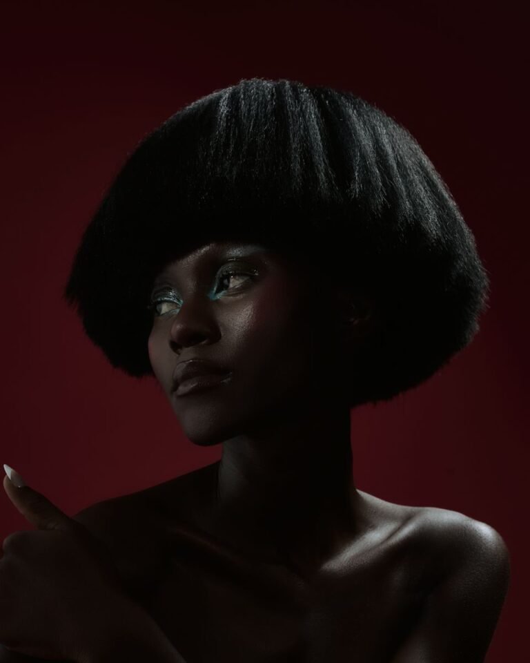 A striking studio portrait of a woman with a bold hairstyle on a dark red background.